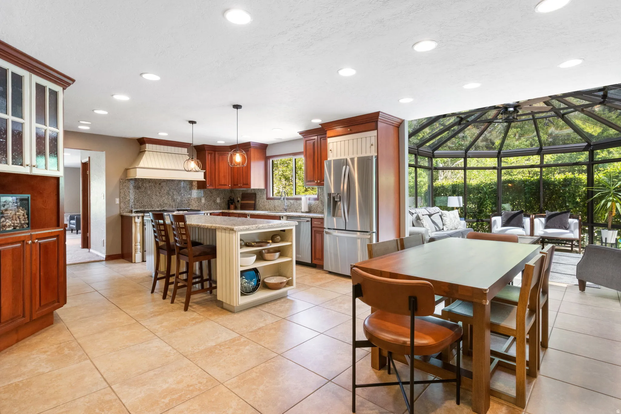Spacious kitchen featuring a large central island with integrated shelving, tile flooring, rich wood cabinetry, stainless steel appliances, and an attached sunroom with a glass ceiling and walls
