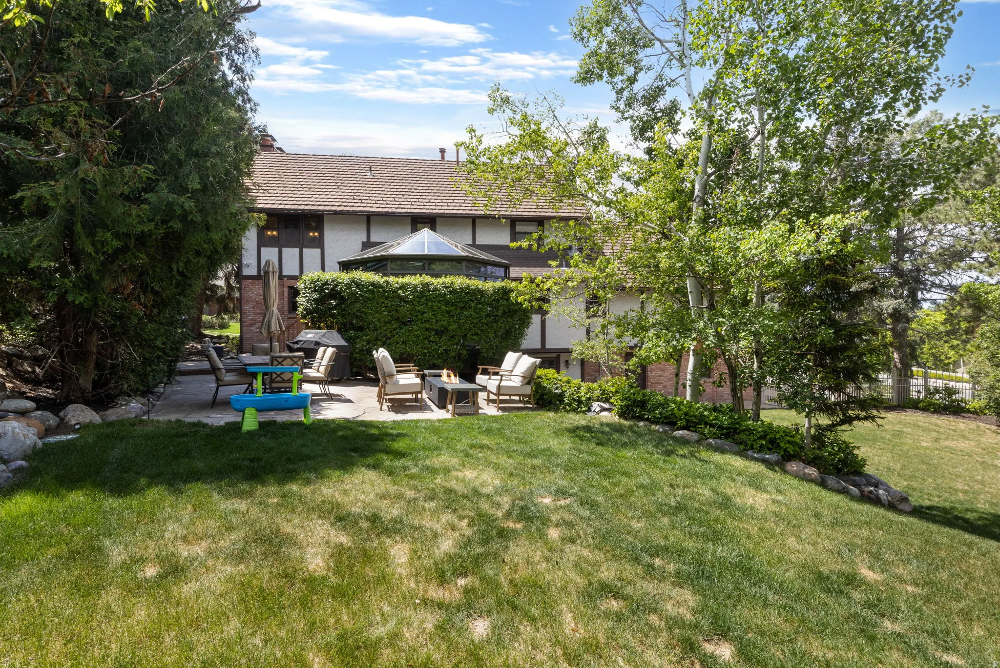 Spacious backyard featuring a manicured lawn, stone retaining walls, mature trees, and a paved patio area