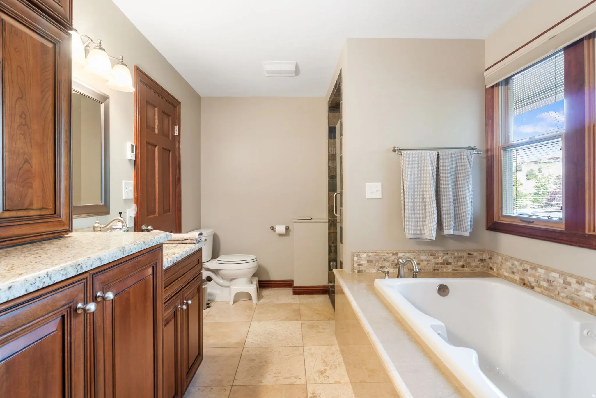 Primary Bathroom.  Bathroom featuring a built-in soaking tub with mosaic tile surround, a separate glass-enclosed shower, wood-finish vanity with a light-toned countertop, and tile flooring