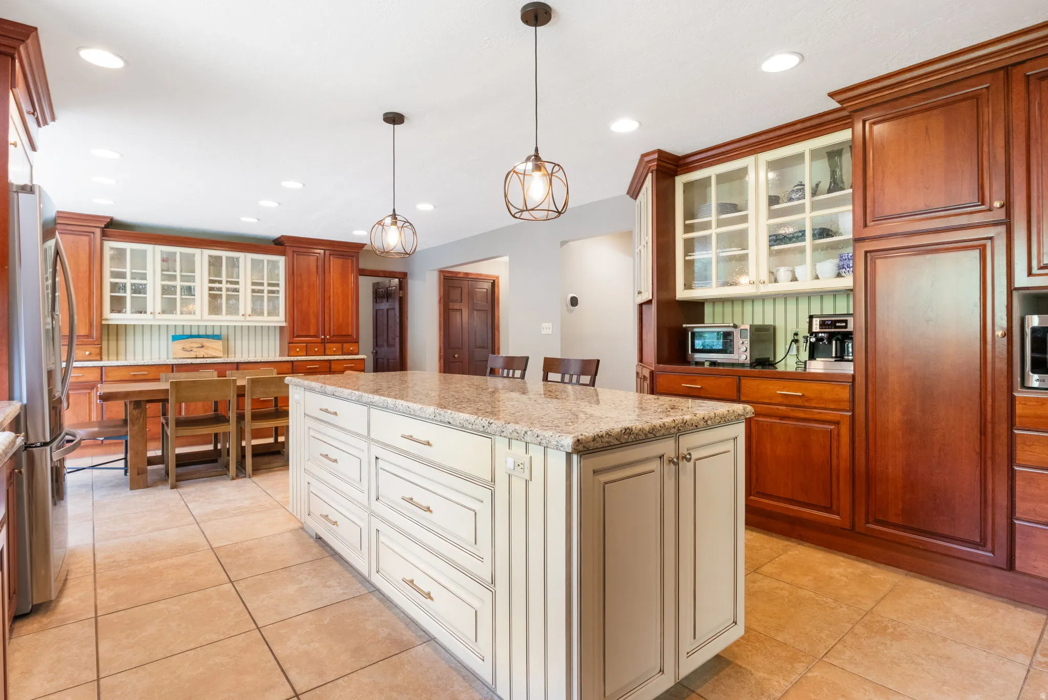 Spacious kitchen featuring a large central island with a light-colored countertop, ceramic tile flooring, extensive wood cabinetry, recessed lighting, and decorative pendant light fixtures
