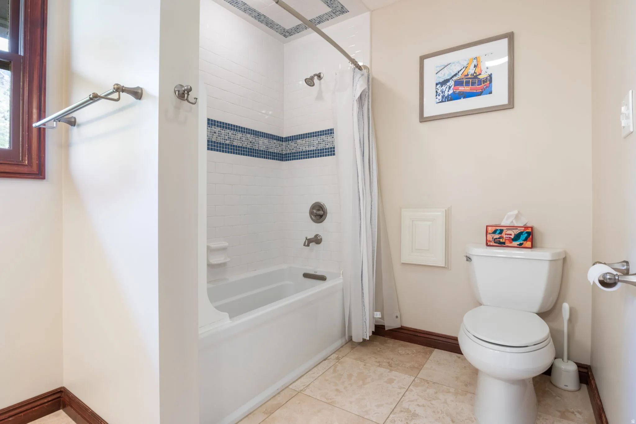 Bathroom featuring a white bathtub with tile surround, decorative blue accent tile, a window with dark wood trim, a toilet, and light-toned floor tiles