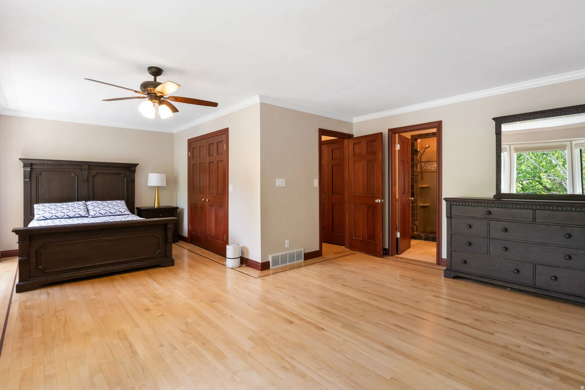 Primary Bathroom with Fireplace.   Spacious room featuring light wood-finish flooring, warm neutral wall tones, white crown molding, a ceiling fan with integrated lighting, and multiple wood-paneled doors