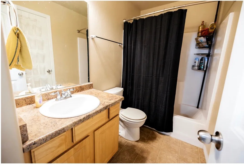 Bathroom featuring a light wood-finish vanity with a white oval sink, a speckled countertop, a large wall mirror, and a white tub with shower