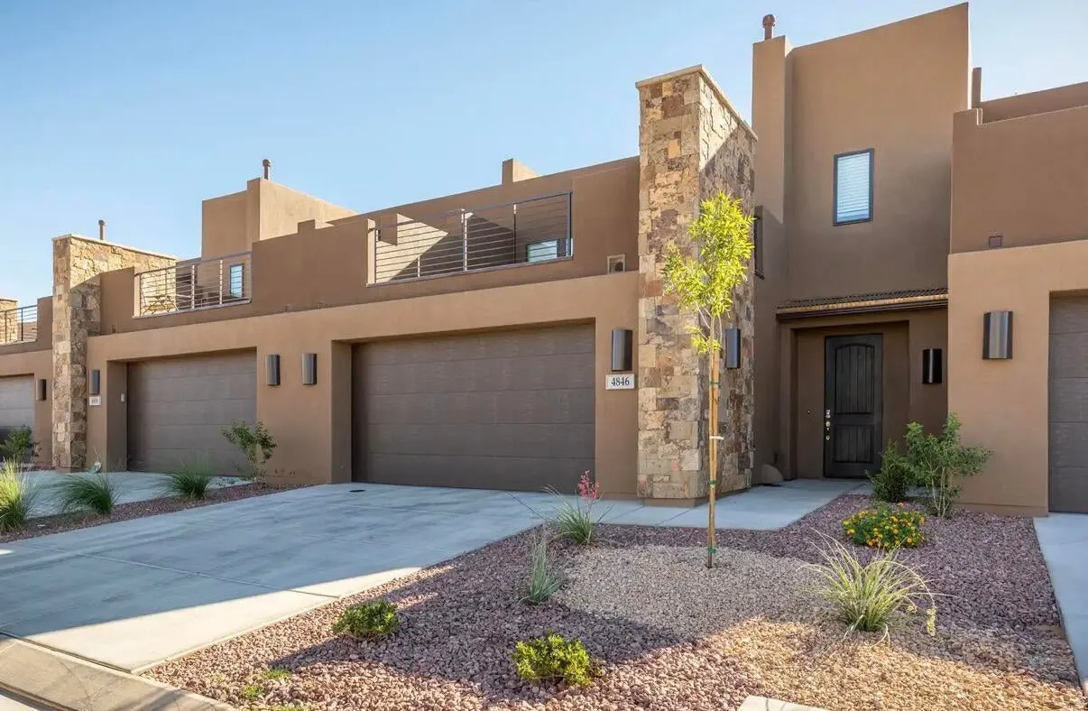 Modern stucco exterior with stone accents, featuring a prominent dark wood entry door, two attached garage doors, and a second-story balcony with horizontal railings