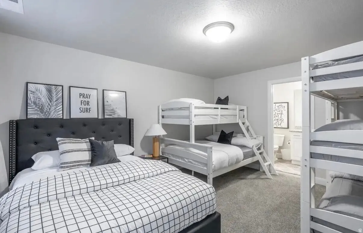 Spacious bedroom featuring neutral wall tones, plush carpet flooring, a flush-mount ceiling light, and access to a full bathroom with a vanity and toilet