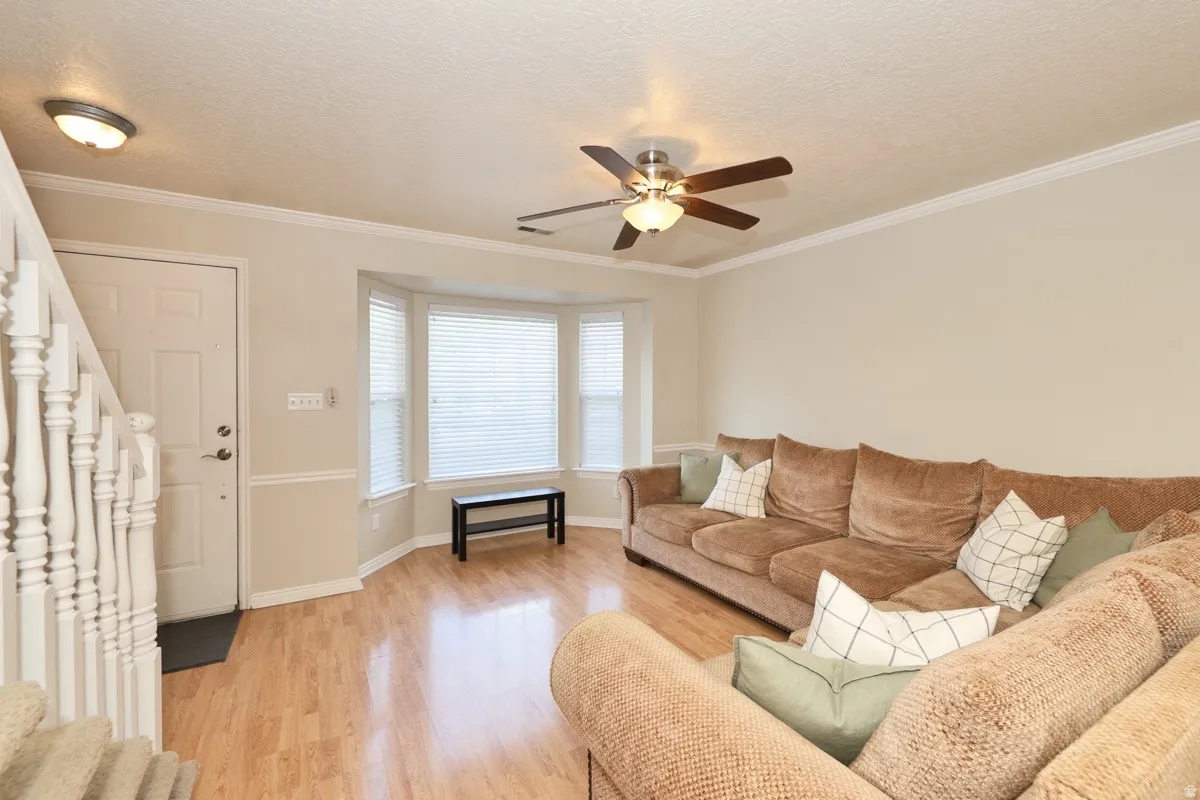 Living area featuring wood-finish flooring, a multi-pane bay window with blinds, crown molding, a ceiling fan with light, and a white entry door