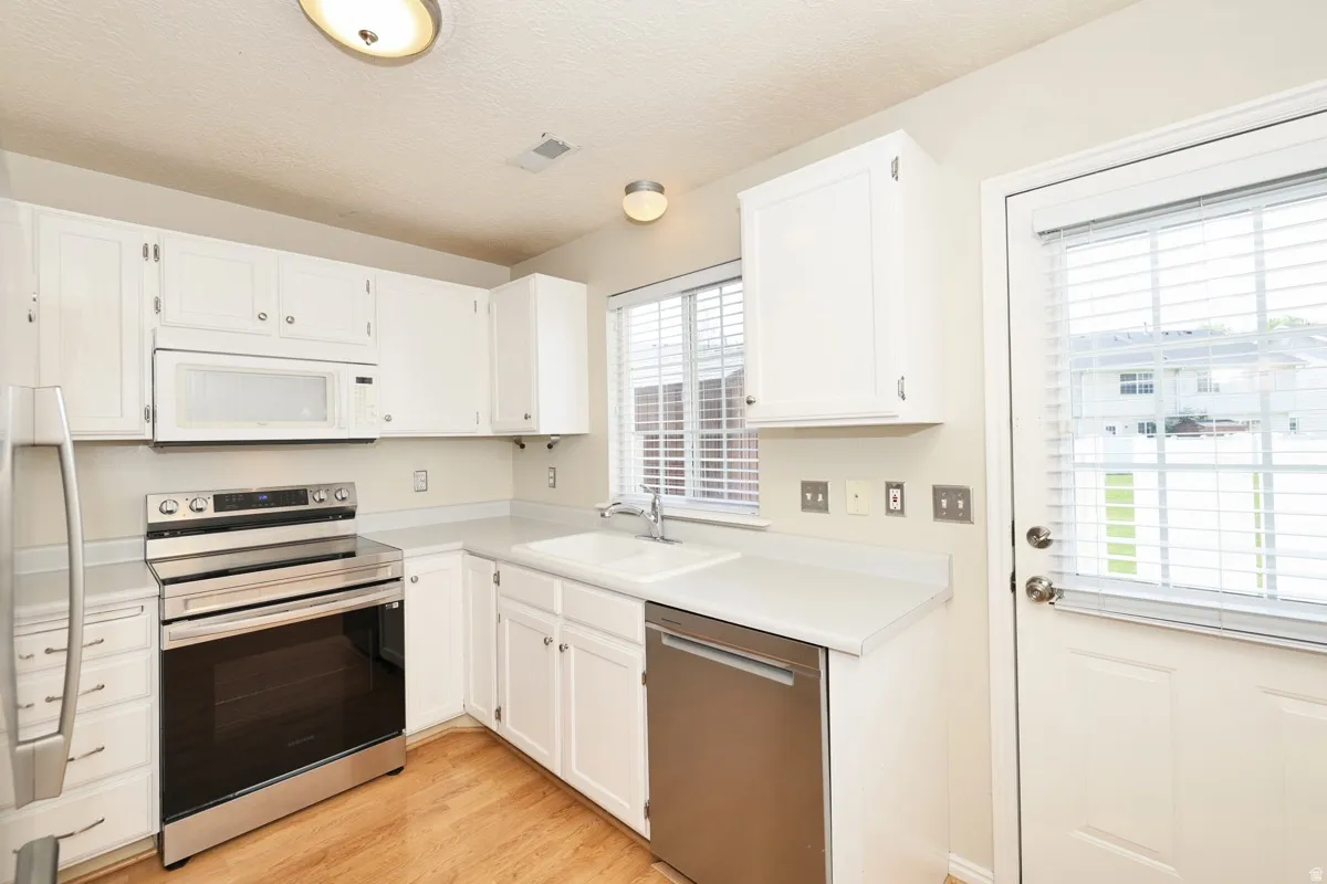 Kitchen featuring white cabinetry, wood-finish flooring, stainless steel appliances, a double basin sink, and a window with blinds