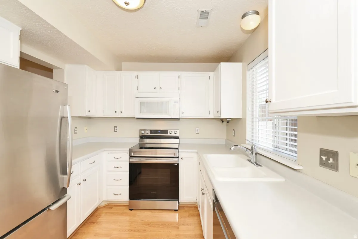 Kitchen featuring white cabinetry, stainless steel appliances, light-toned countertops, a dual-basin sink with chrome faucet, and wood-finish flooring