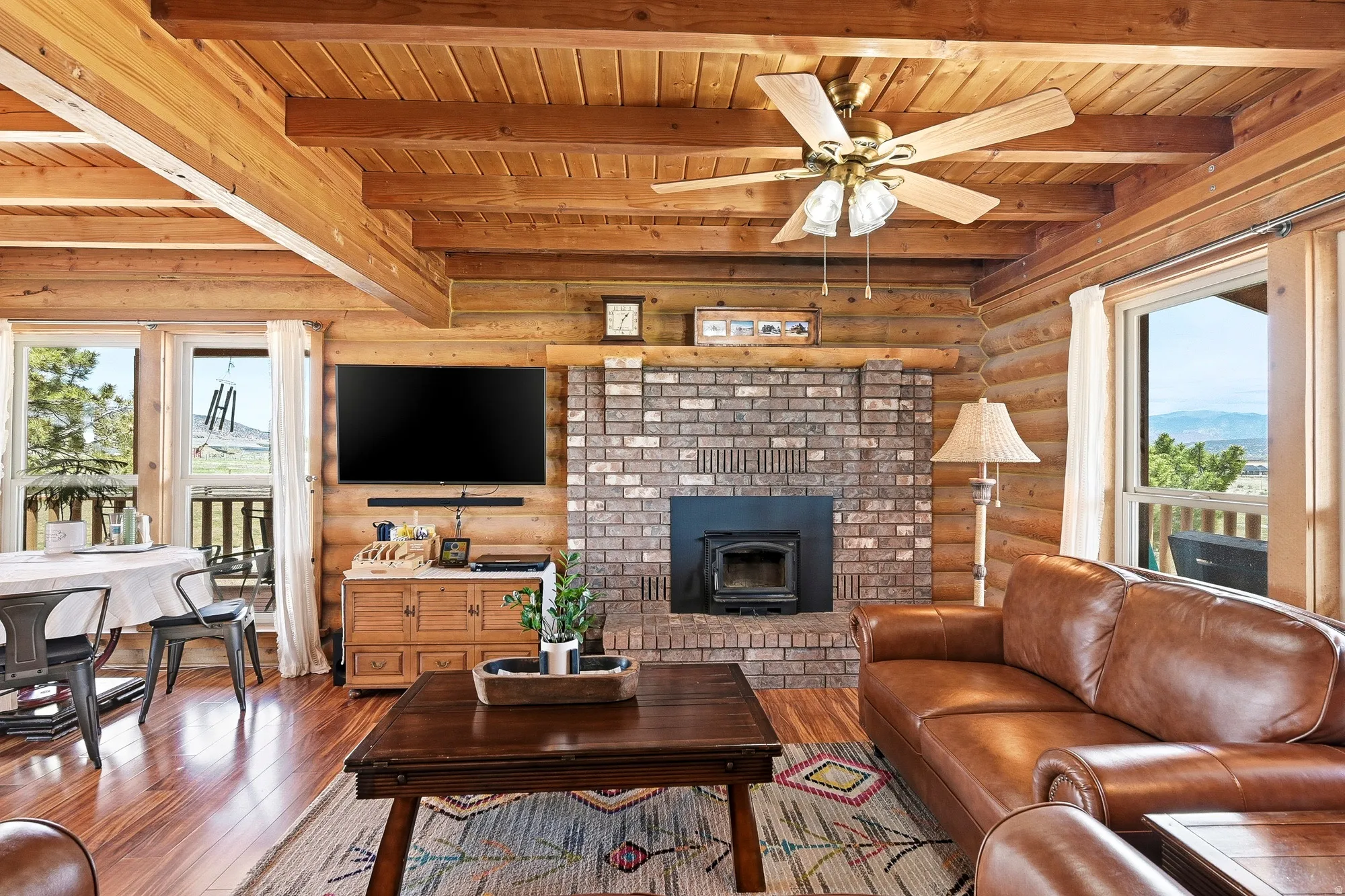 Living area featuring log wall construction, exposed beam ceiling, brick fireplace with wood stove insert, wood-finish flooring, and multiple windows