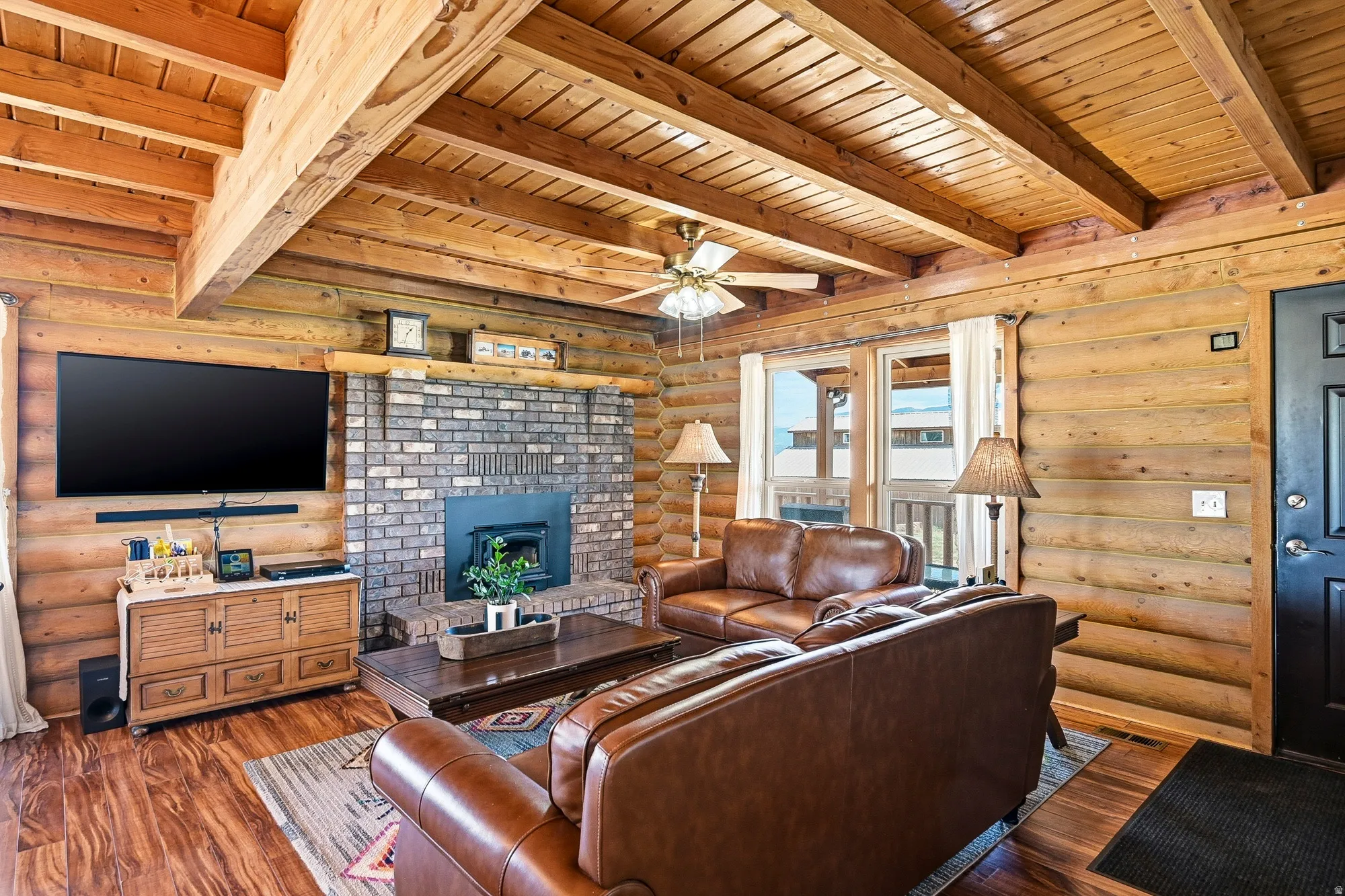 Living area featuring exposed wood beam ceiling, wood-finish flooring, a brick fireplace with mantel, log walls, and multiple windows