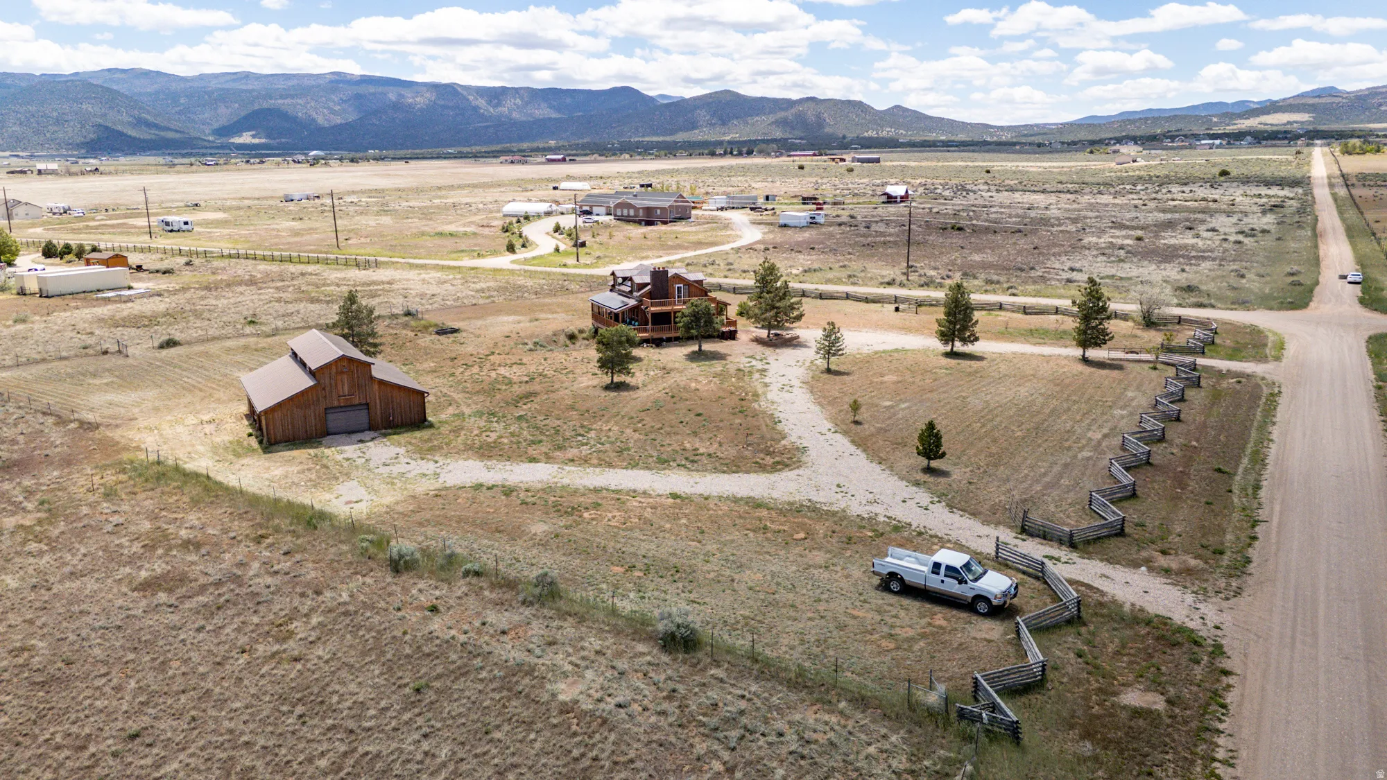 Expansive property featuring a multi-level residence with exterior wood siding and multiple balconies, a separate barn structure, a meandering gravel driveway, a decorative wooden fence, and mature trees on a vast rural landscape