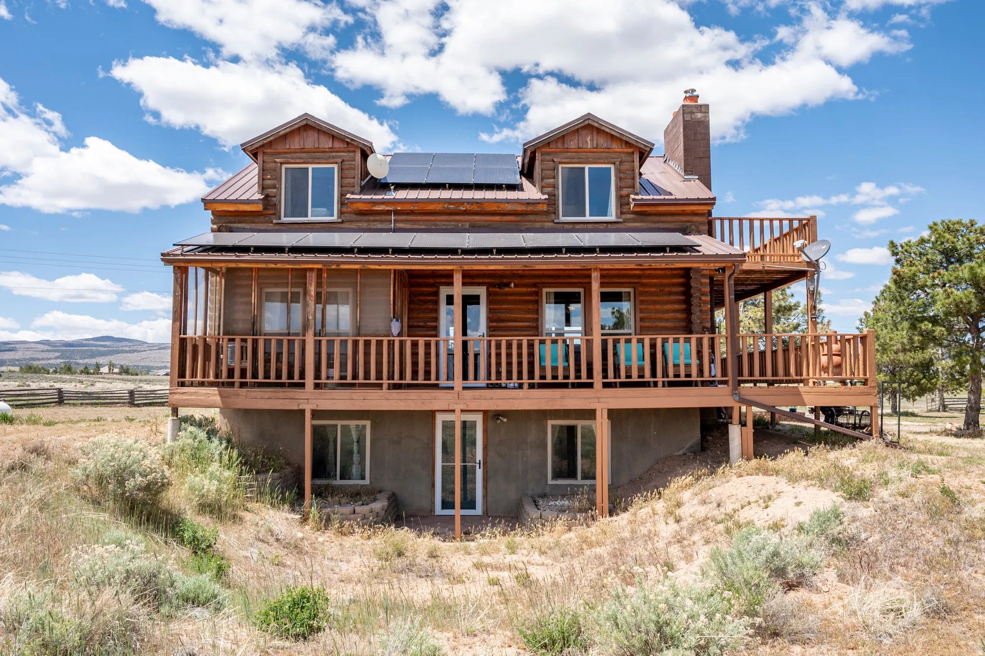 Log-sided residence featuring a full-width covered porch, an upper-level deck, a brown metal roof, dormer windows, and visible solar panels