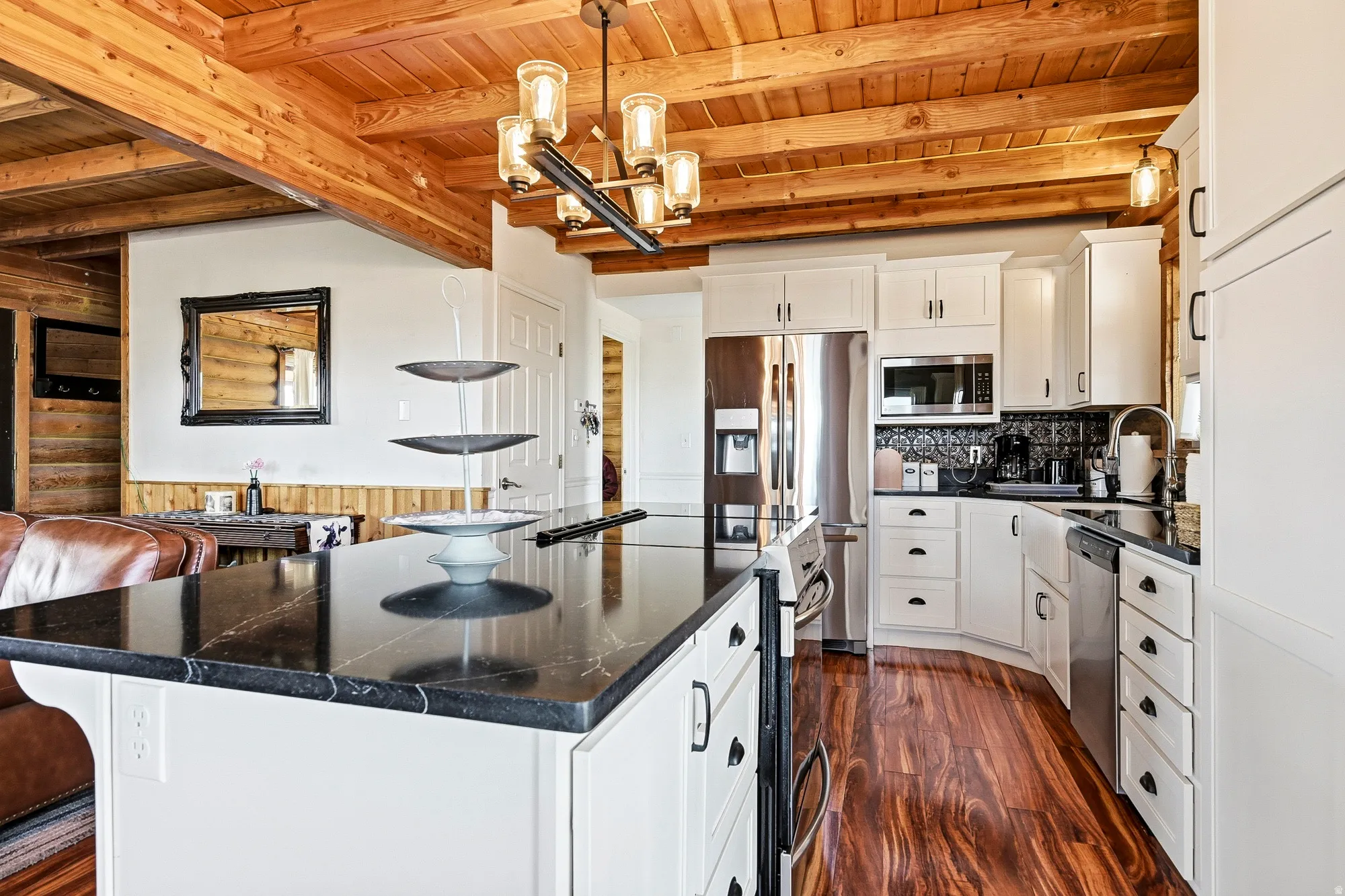 Kitchen island with a dark stone countertop, white cabinetry with dark hardware, stainless steel appliances, wood-finish flooring, and a wood-beamed ceiling