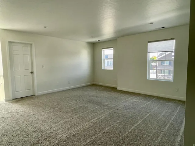 Spacious carpeted room featuring two windows with roller shades, a six-panel interior door, and recessed ceiling lighting
