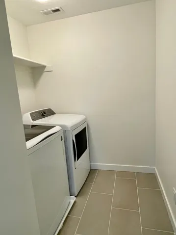 Laundry area featuring neutral wall tones, a built-in storage shelf, and light-toned rectangular tile flooring