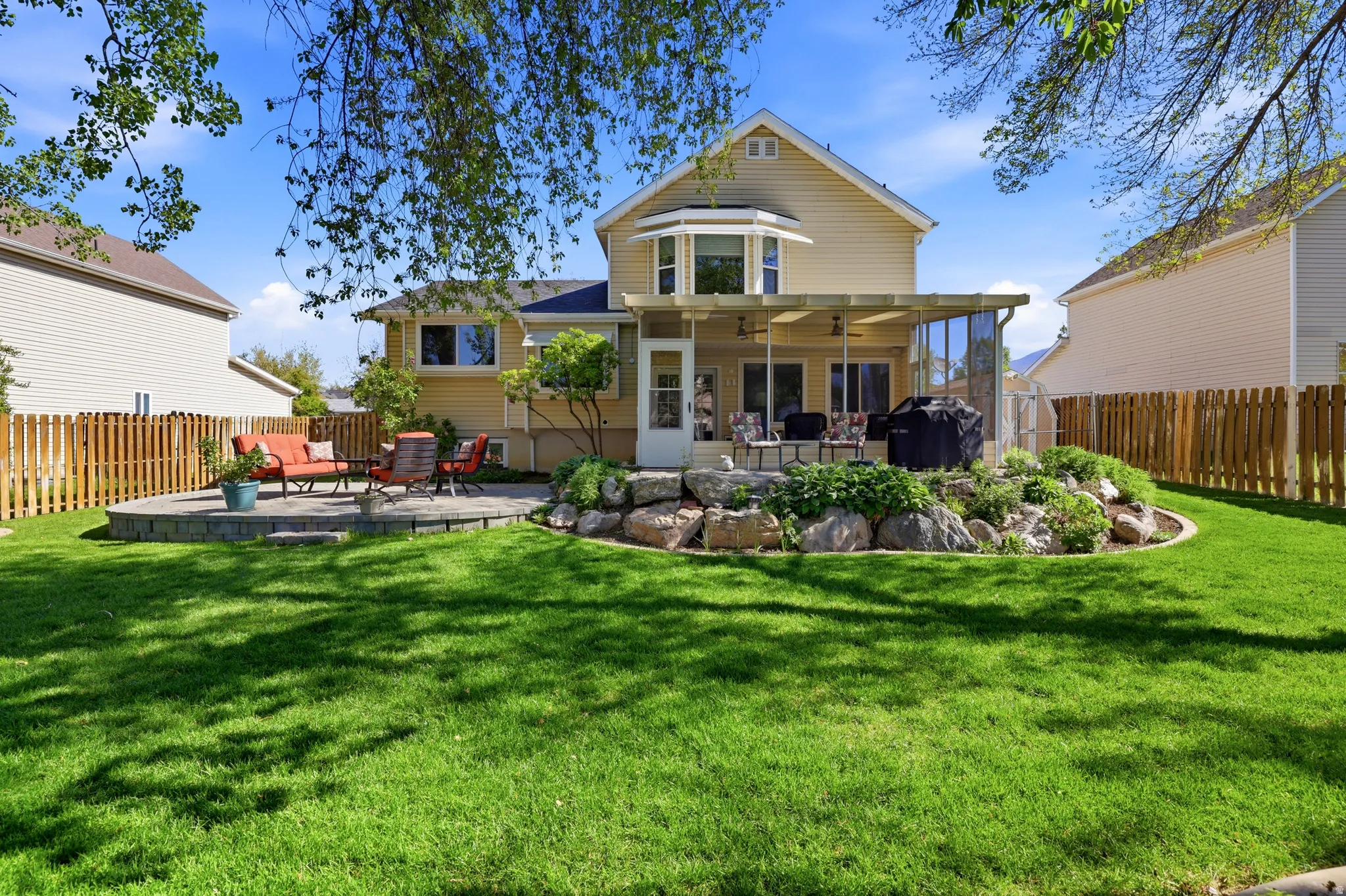 Expansive backyard featuring a large green lawn, two distinct patio areas, a covered pergola, a rock-lined garden bed, and wood privacy fencing