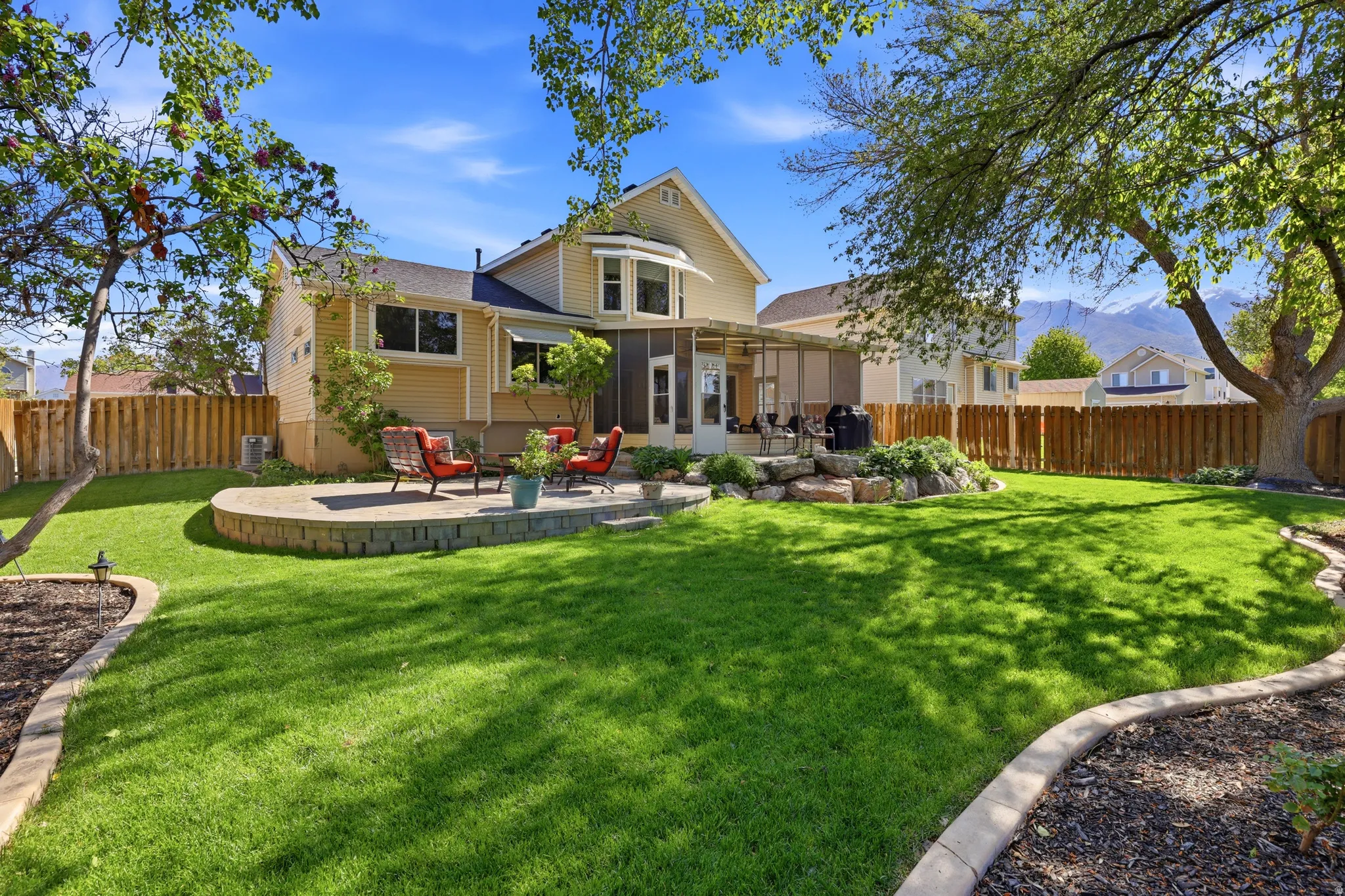 Expansive backyard featuring a manicured lawn, a raised stone-paver patio, a screened porch, mature perimeter trees, and a mountain backdrop