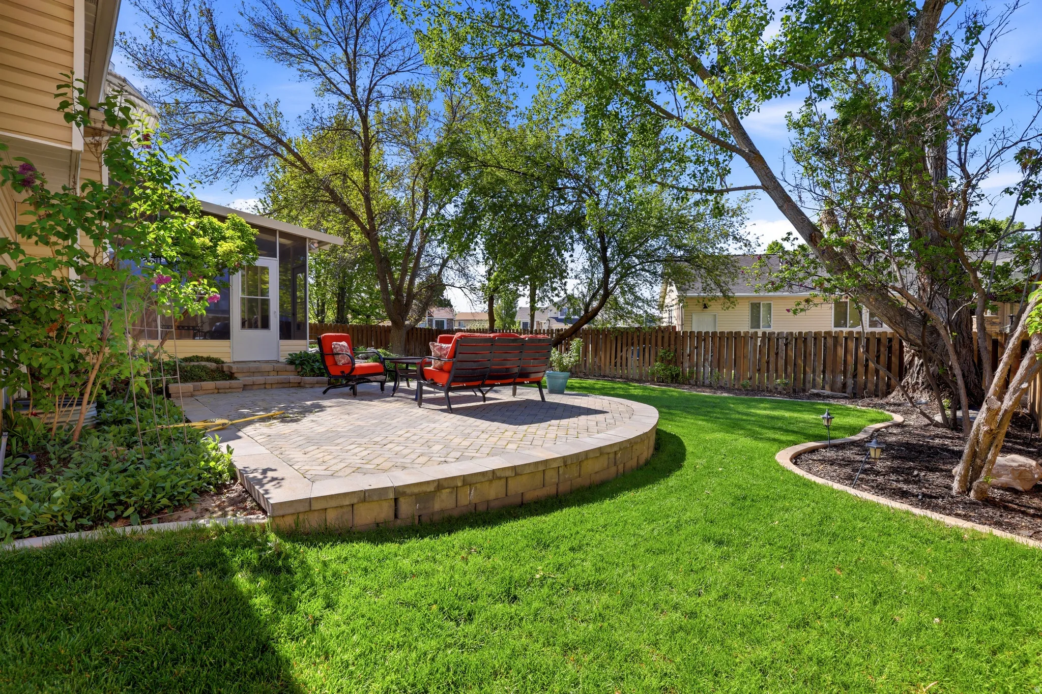 Expansive backyard featuring a curved paver patio with a low retaining wall, a screened-in porch with a white door, and mature trees providing natural shade