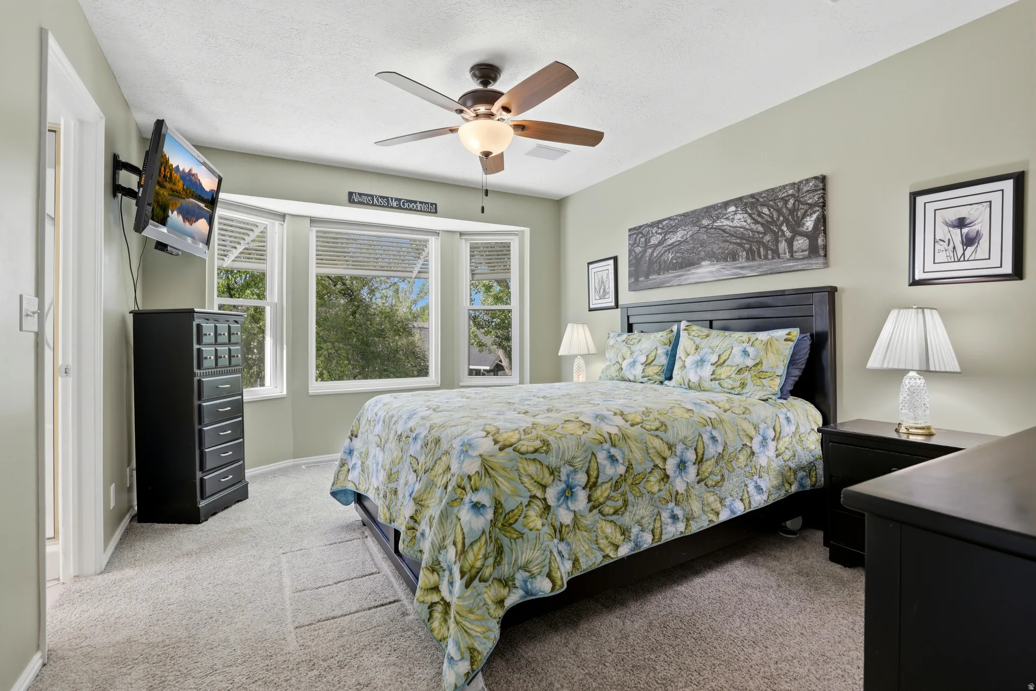 Master Carpeted room featuring a ceiling fan with light fixture, bay windows with blinds, a wall-mounted television bracket, and a paneled bed frame