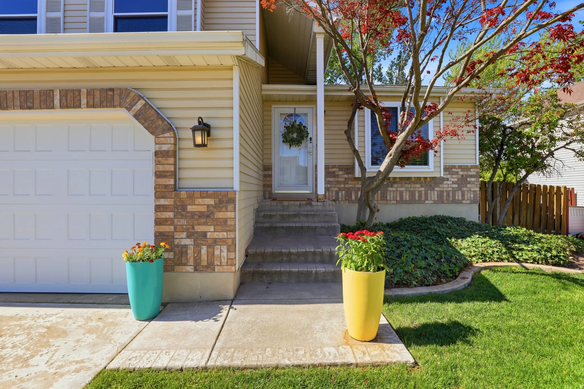 Exterior featuring tan siding, brick accents, a two-car garage, established landscaping, and a concrete walkway