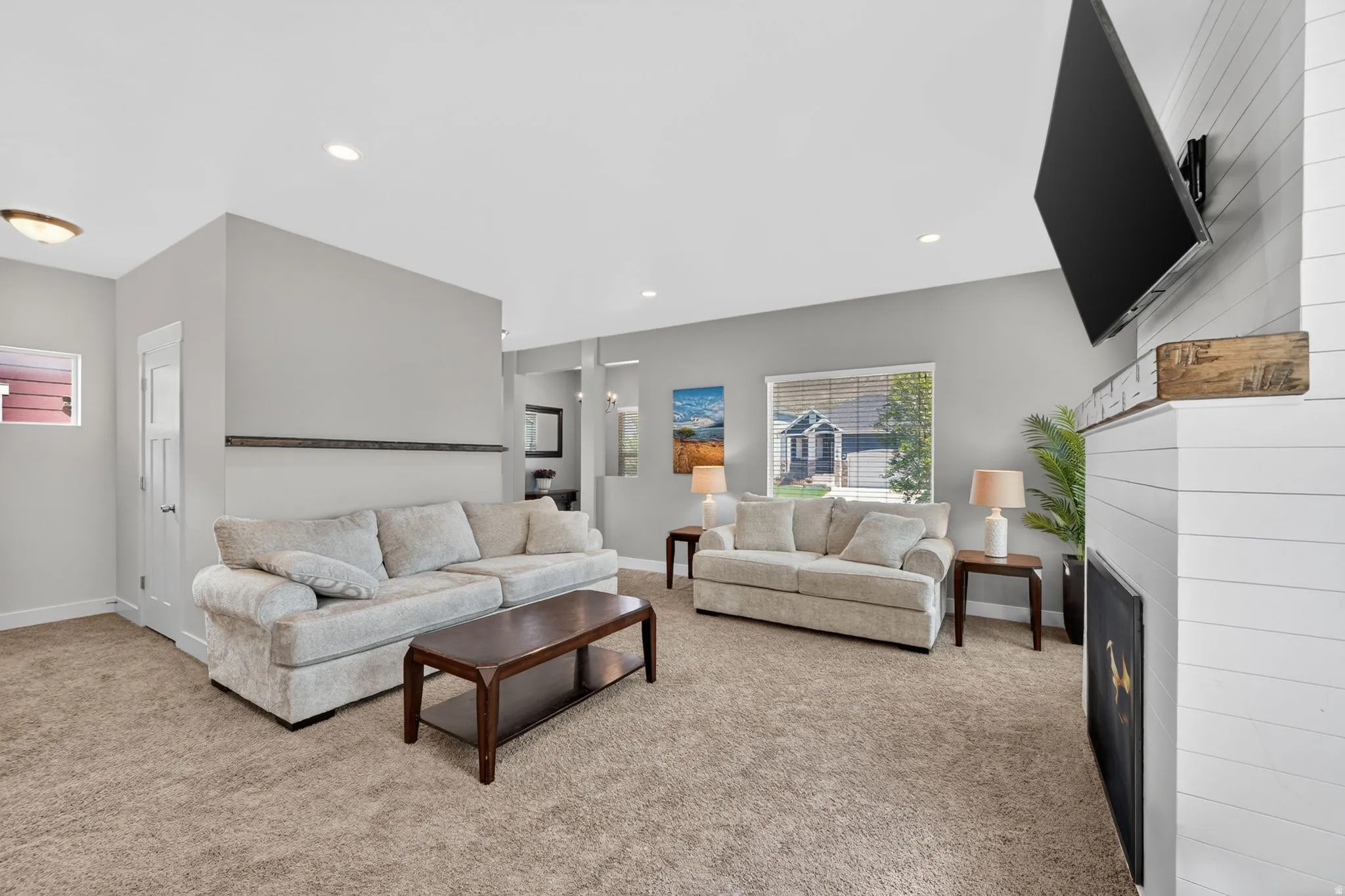 Spacious living area featuring neutral carpeting, light gray walls, recessed lighting, a fireplace with a wood-finish mantel and shiplap-style surround, and a large window providing ample natural light