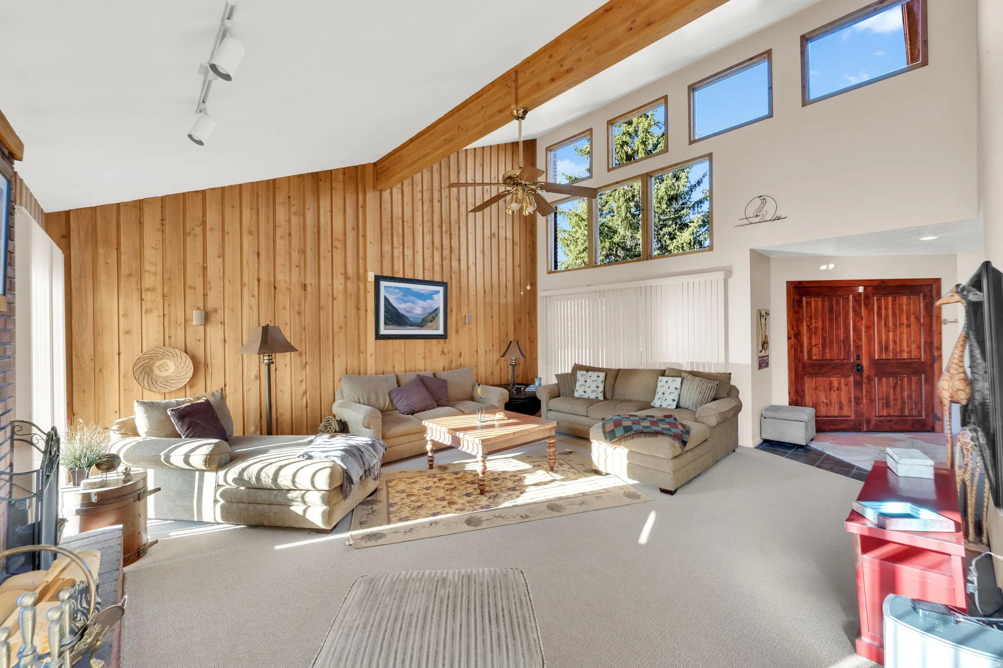 Spacious living area featuring vaulted ceilings with exposed wood beams, a full wall of natural wood paneling, and a floor-to-ceiling window arrangement including transom windows