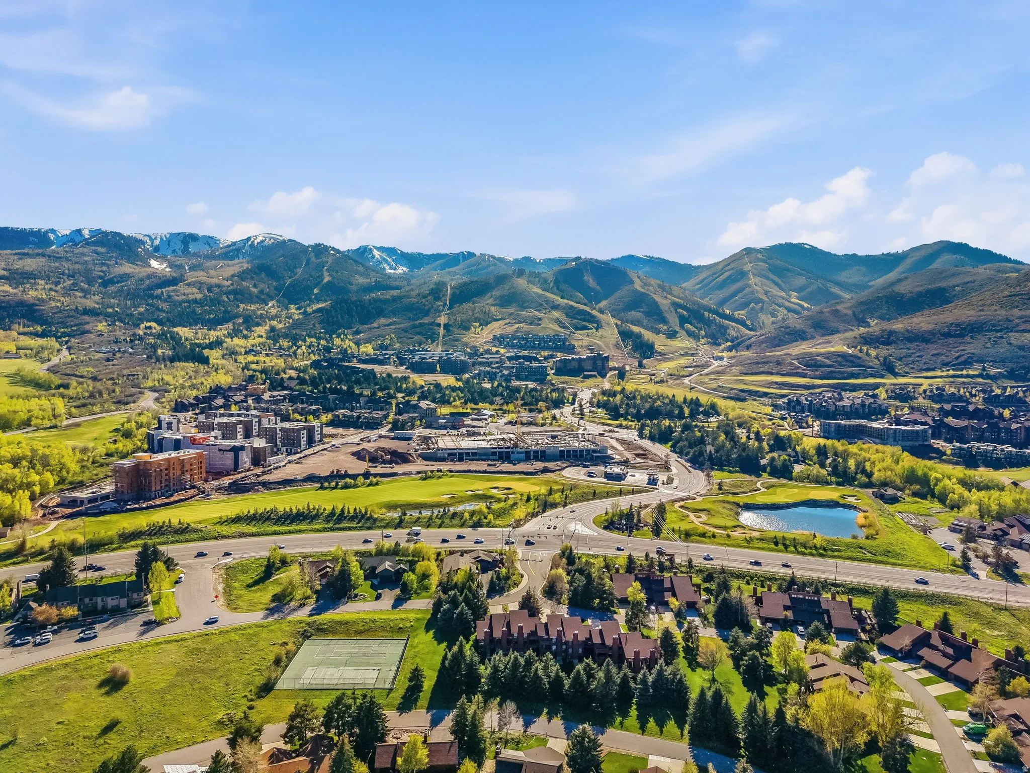 Expansive mountain landscape featuring snow-capped peaks, verdant hillsides, a substantial body of water, and a tennis court