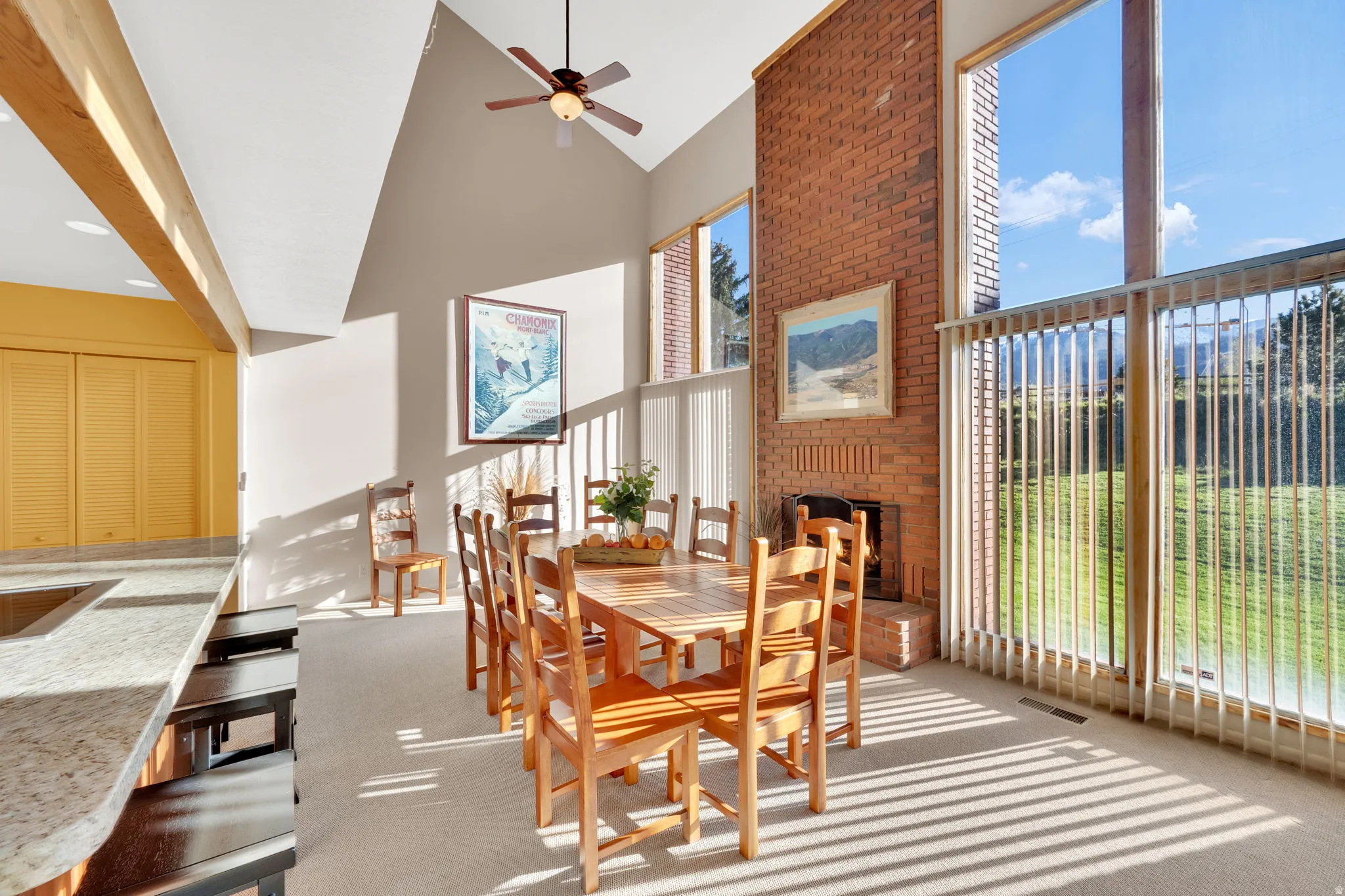 Vaulted ceiling dining area featuring a full-height brick fireplace, a ceiling fan, and extensive window walls with vertical blinds