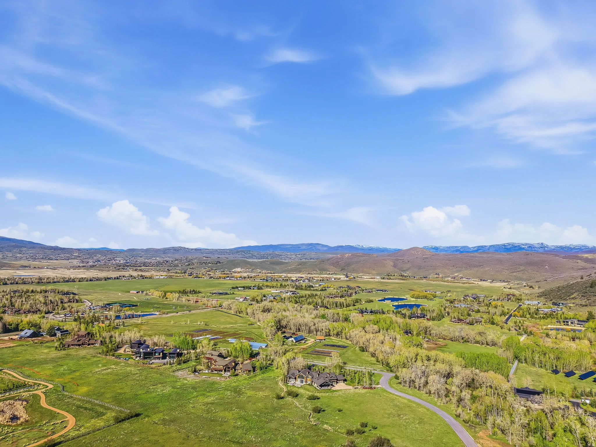 Expansive rural landscape featuring rolling green hills, scattered mature trees, distant mountains, and a clear blue sky