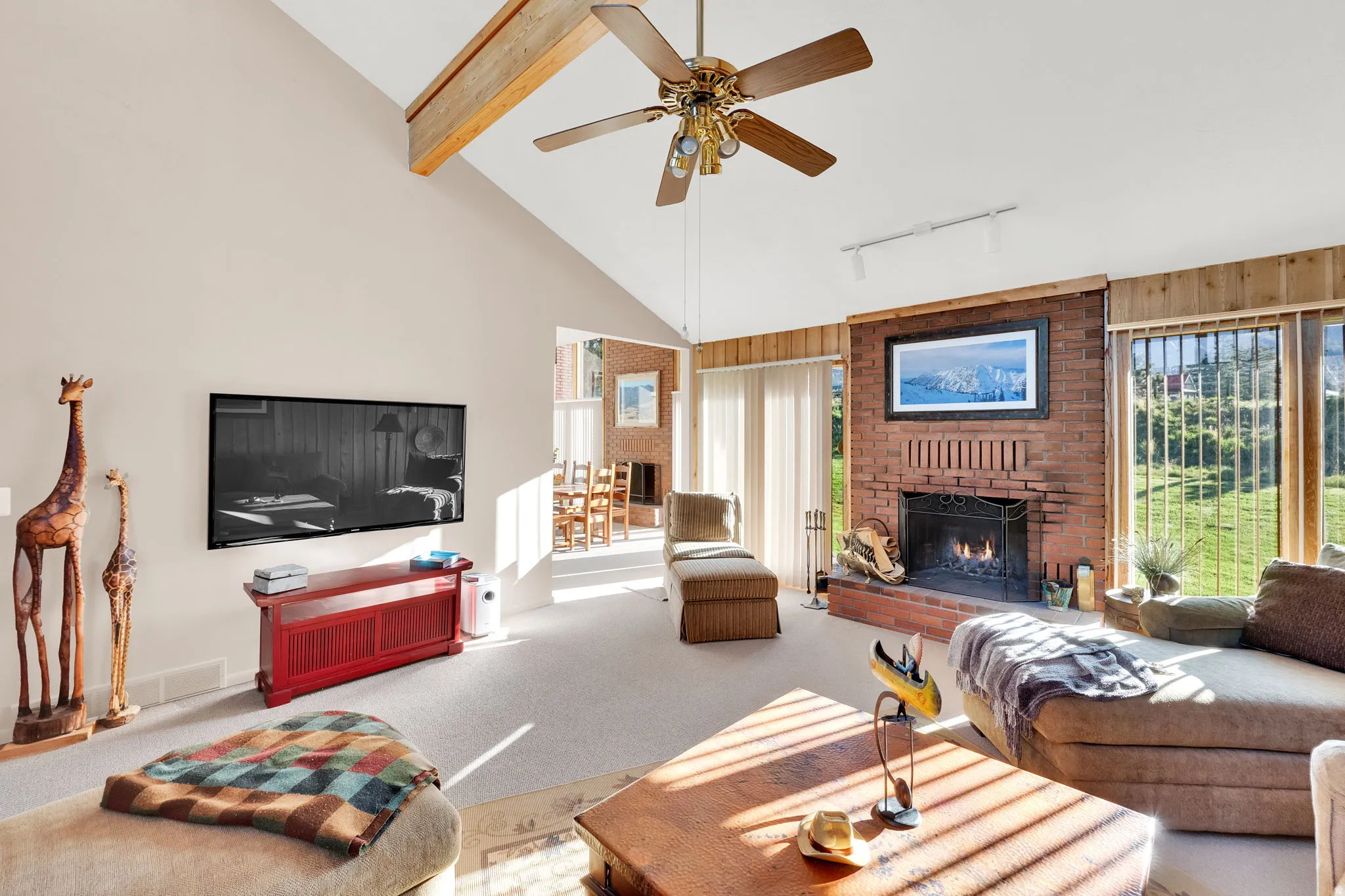 Spacious living area featuring a brick fireplace, vaulted ceiling with exposed wood beam, ceiling fan, and extensive window-lined walls