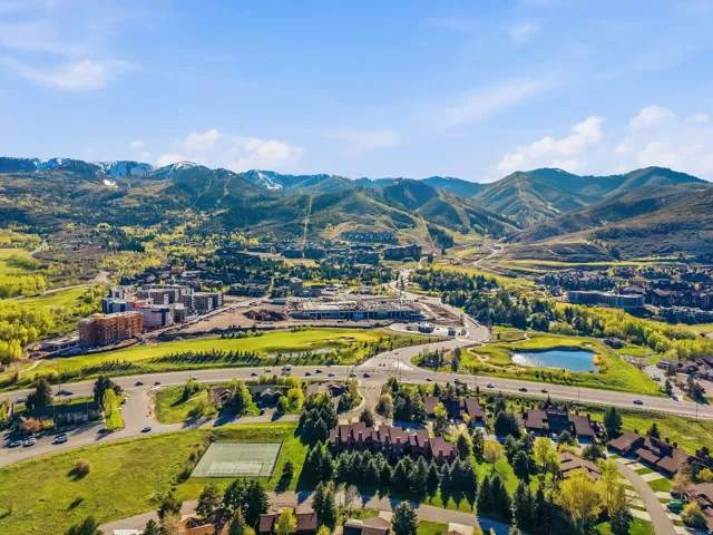 Expansive mountain landscape featuring snow-capped peaks, verdant hillsides, a substantial body of water, and a tennis court
