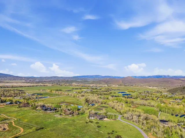 Expansive rural landscape featuring rolling green hills, scattered mature trees, distant mountains, and a clear blue sky