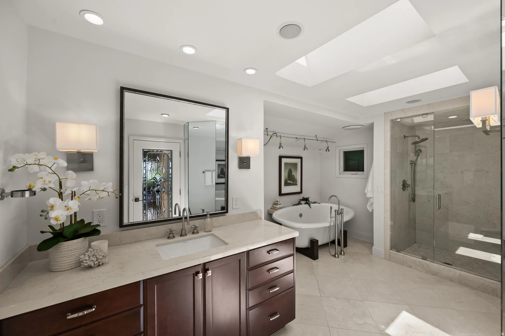 Spacious bathroom featuring a freestanding soaking tub, glass-enclosed shower with multiple showerheads, dual skylights, a dark wood-finish vanity with an undermount sink, and light-toned countertop