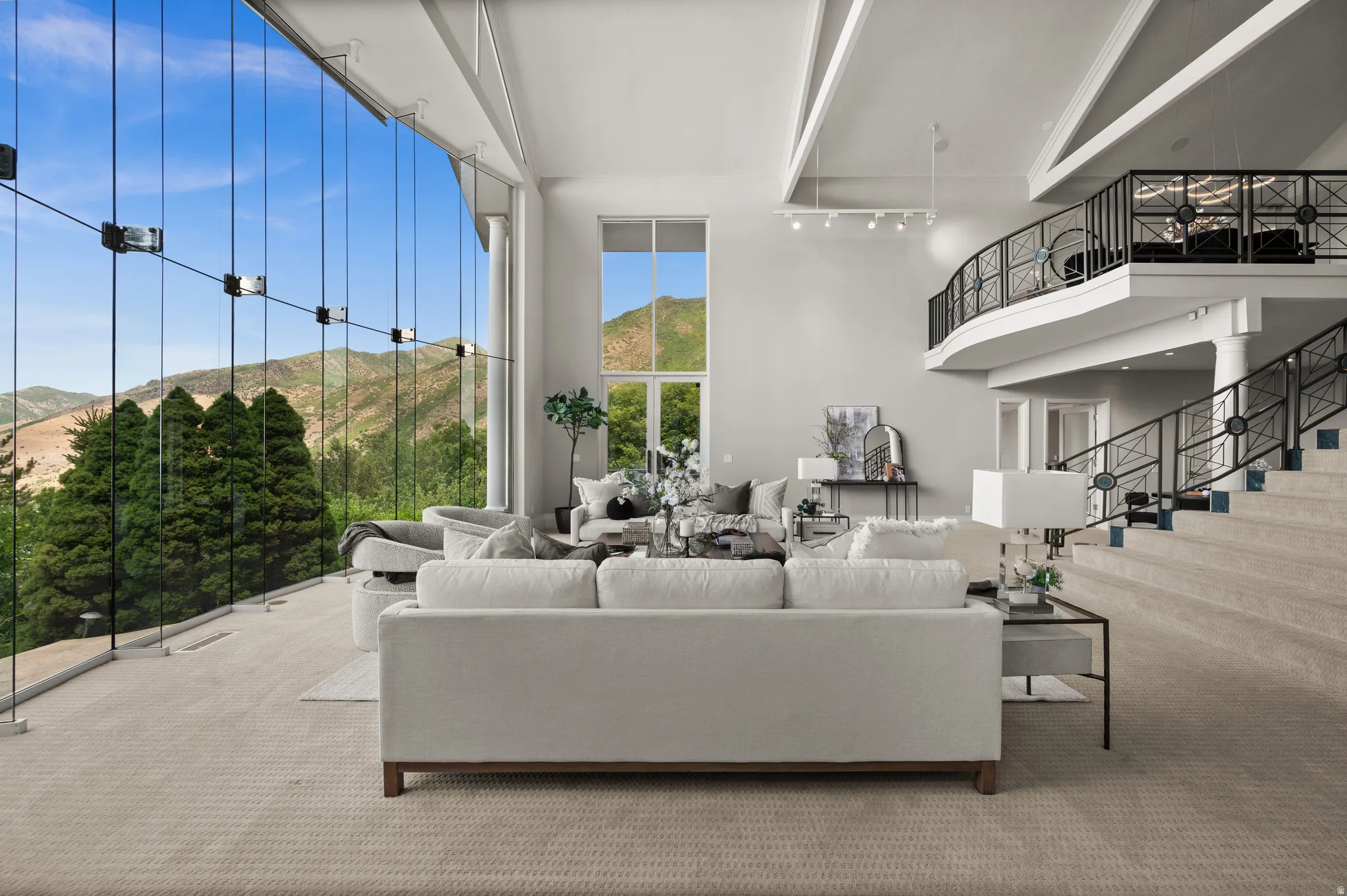 Spacious living area featuring floor-to-ceiling glass panel walls, vaulted ceilings with exposed beams, a grand staircase with decorative metal railings, and an upper-level balcony