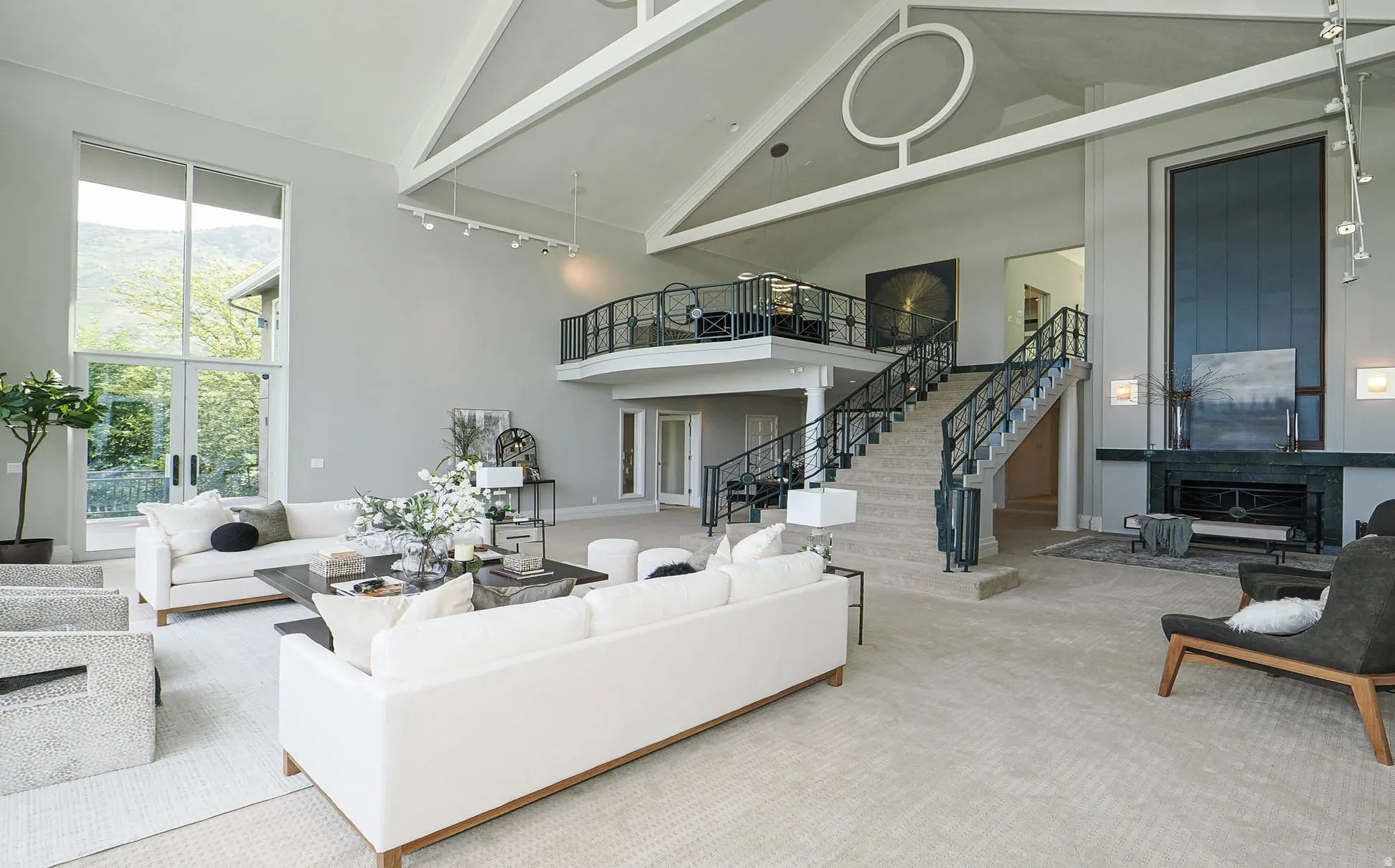 Grand living space featuring soaring vaulted ceilings, a prominent staircase with wrought iron railings, an upper-level balcony, a built-in fireplace with a stone surround, and large glass paneled doors