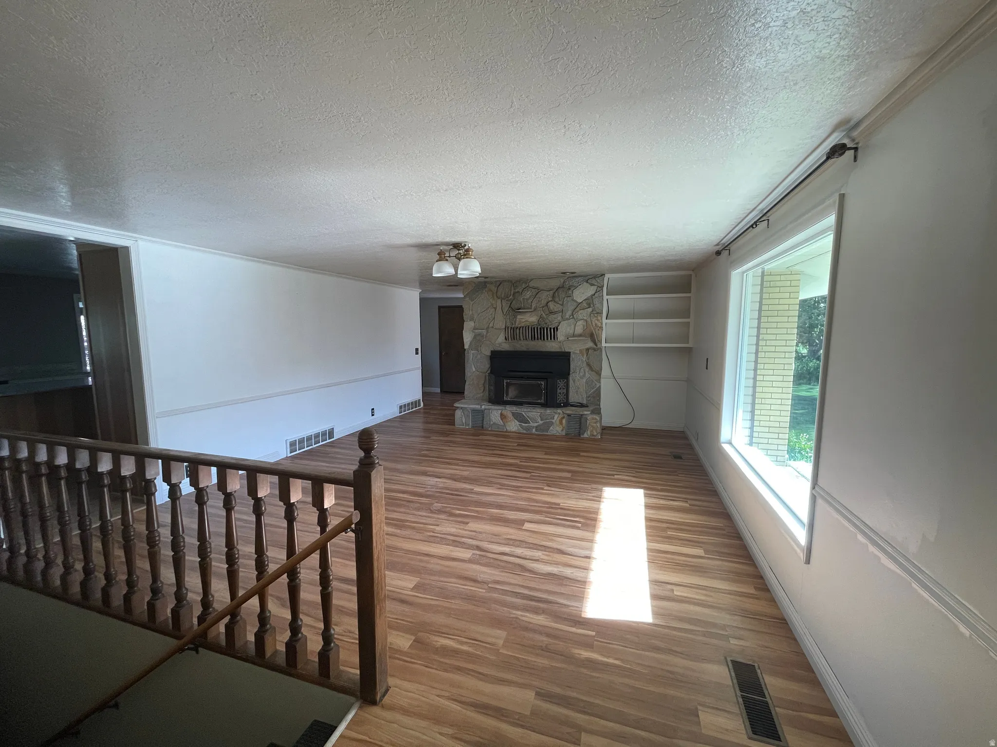 Spacious living area featuring wood-finish flooring, a stone fireplace with hearth, a large picture window, built-in shelving, and a classic wooden staircase railing