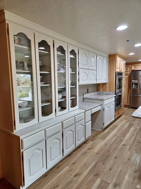 Kitchen featuring white cabinetry with glass display doors, recessed lighting, wood-finish flooring, light-toned countertops, and built-in double ovens