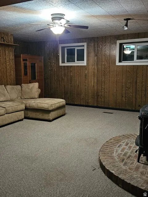 Carpeted room featuring wood paneling, two windows with white trim, a ceiling fan with integrated lighting, and a brick hearth