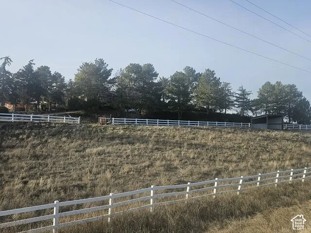 Sloping terrain featuring a white multi-rail fence, mature trees, and a utility building