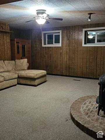 Carpeted room featuring wood paneling, two windows with white trim, a ceiling fan with integrated lighting, and a brick hearth