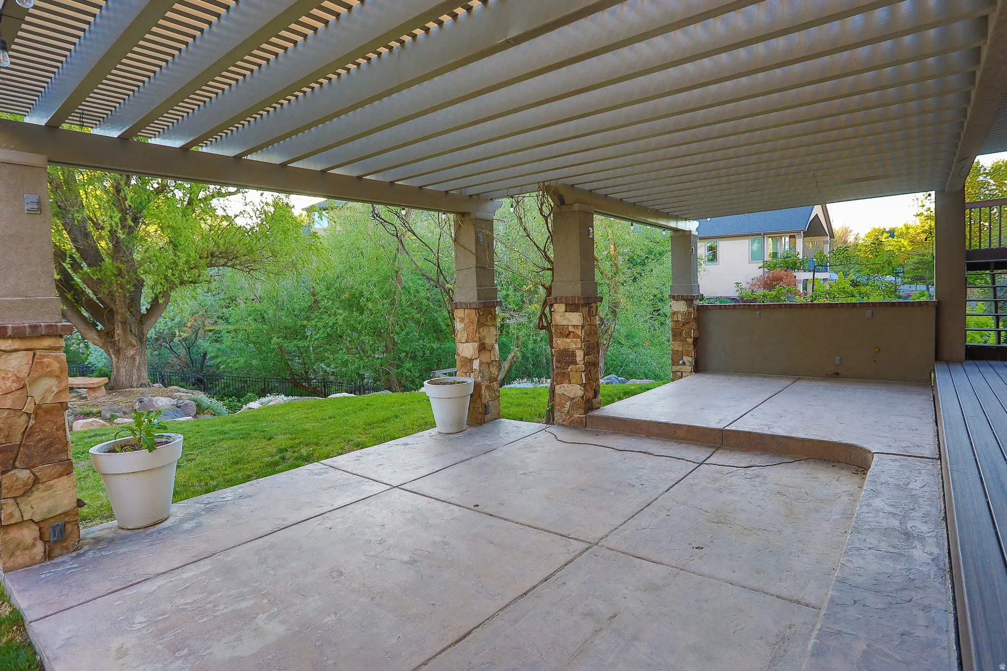 Expansive outdoor patio featuring a slatted pergola, stone-clad support columns, a concrete slab, and a composite deck extension