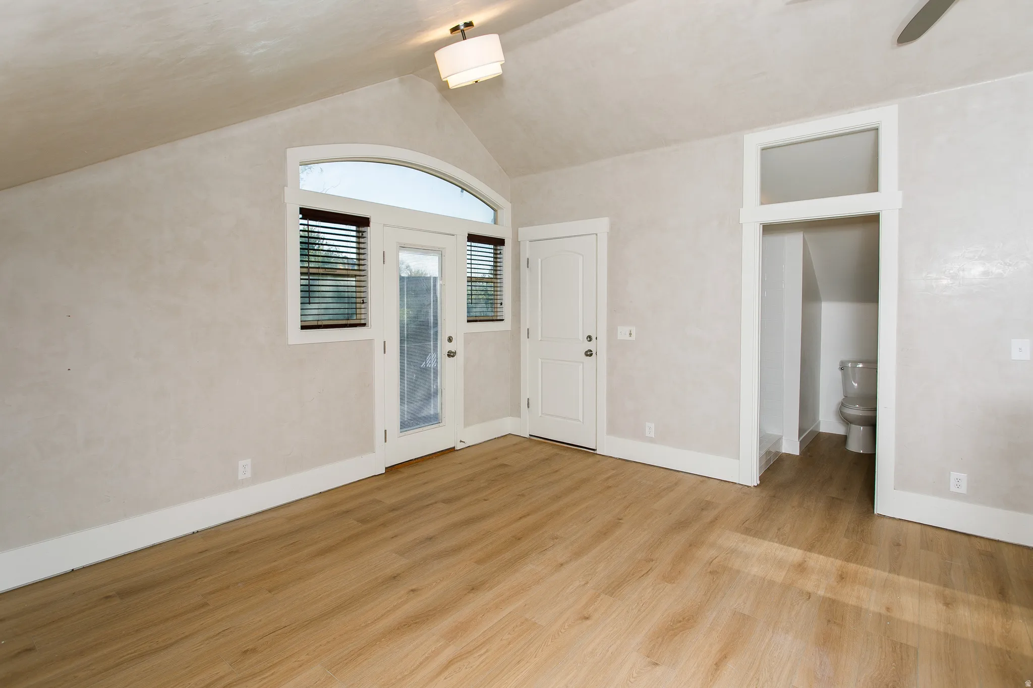 Vaulted ceiling room with wood-finish flooring, a white glass-paneled door, and an arched transom window