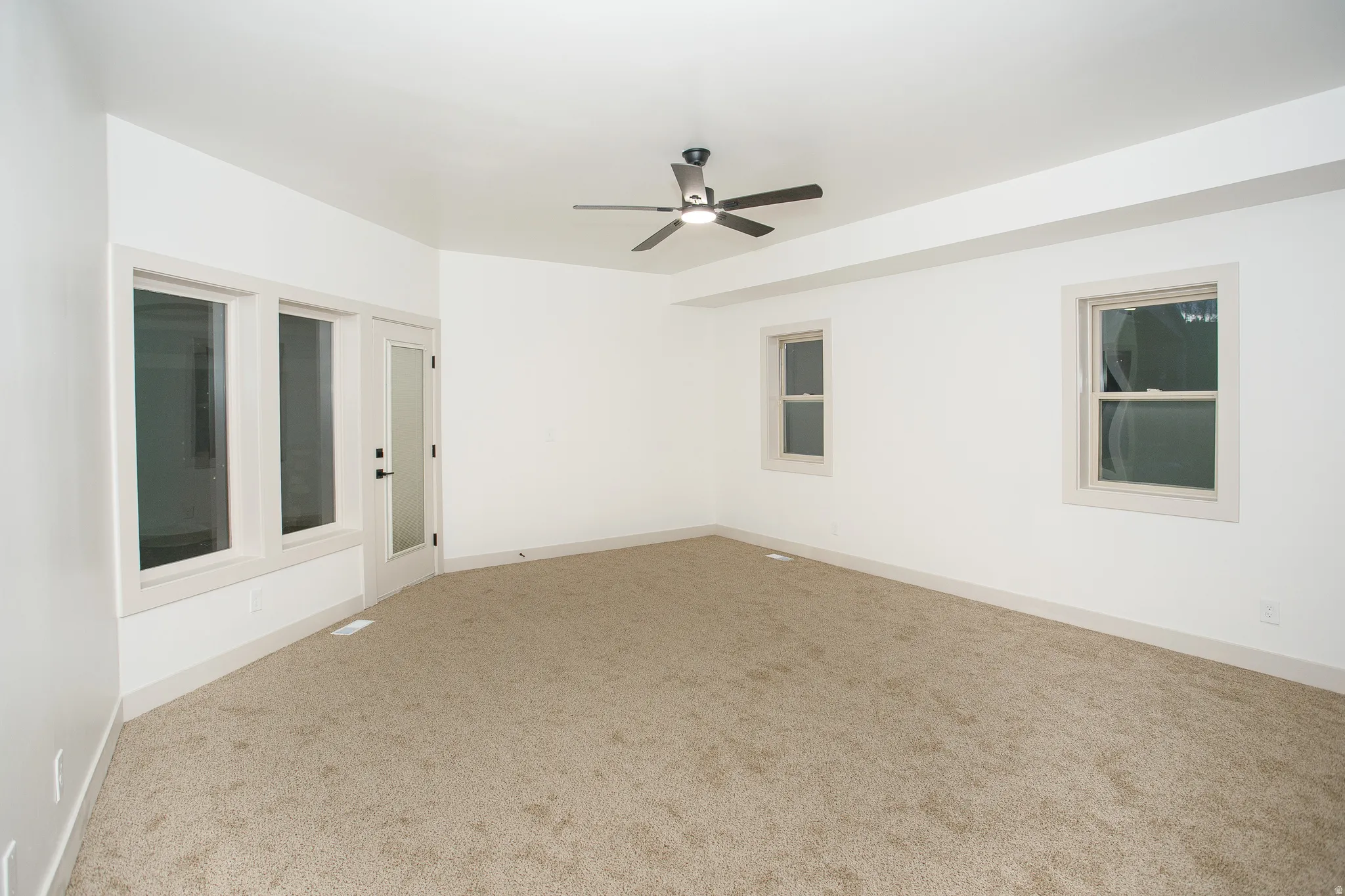 Spacious room with plush carpeting, a contemporary ceiling fan, multiple windows for natural illumination, a glass-paneled exterior door, and a tray ceiling detail