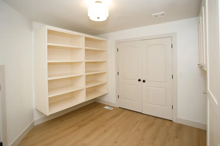 Built-in shelving unit with multiple tiers, light wood-finish flooring, white double doors with black hardware, and overhead lighting fixture
