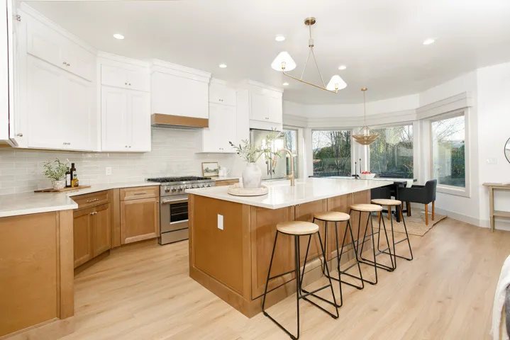 Open-concept kitchen featuring a large central island with seating, a professional-grade stainless steel range, light wood-finish flooring, two-tone cabinetry with white upper and wood-tone lower cabinets, and a bay window dining area