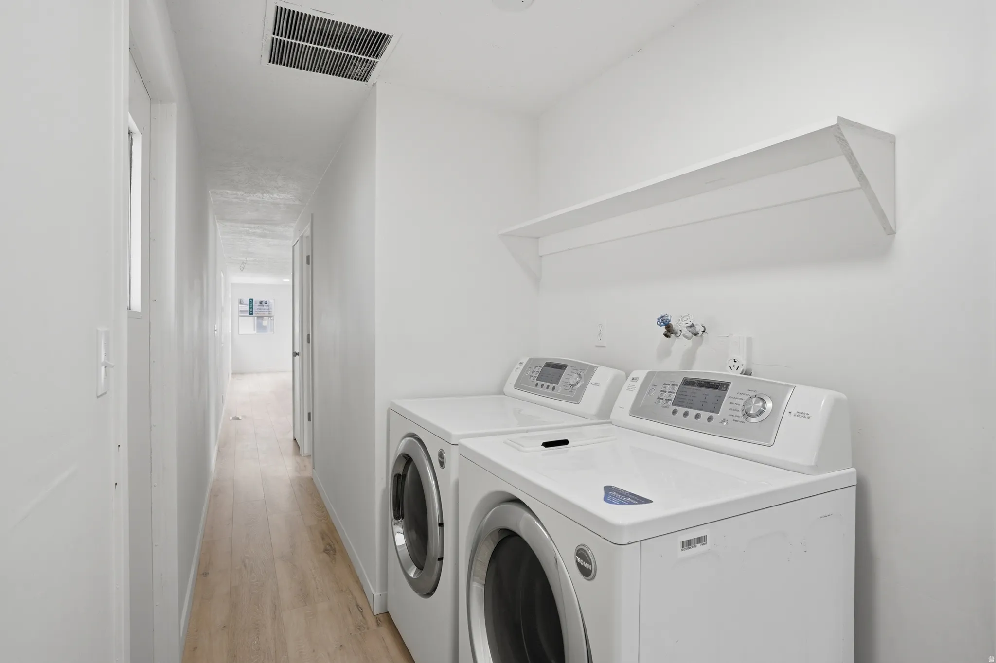 Dedicated laundry area featuring a white wall-mounted shelf, light wood-finish flooring, and crisp white walls