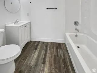 Bathroom featuring wood-finish flooring, a white vanity with integrated sink, circular wall mirror, toilet, and a white bathtub with shower fixtures