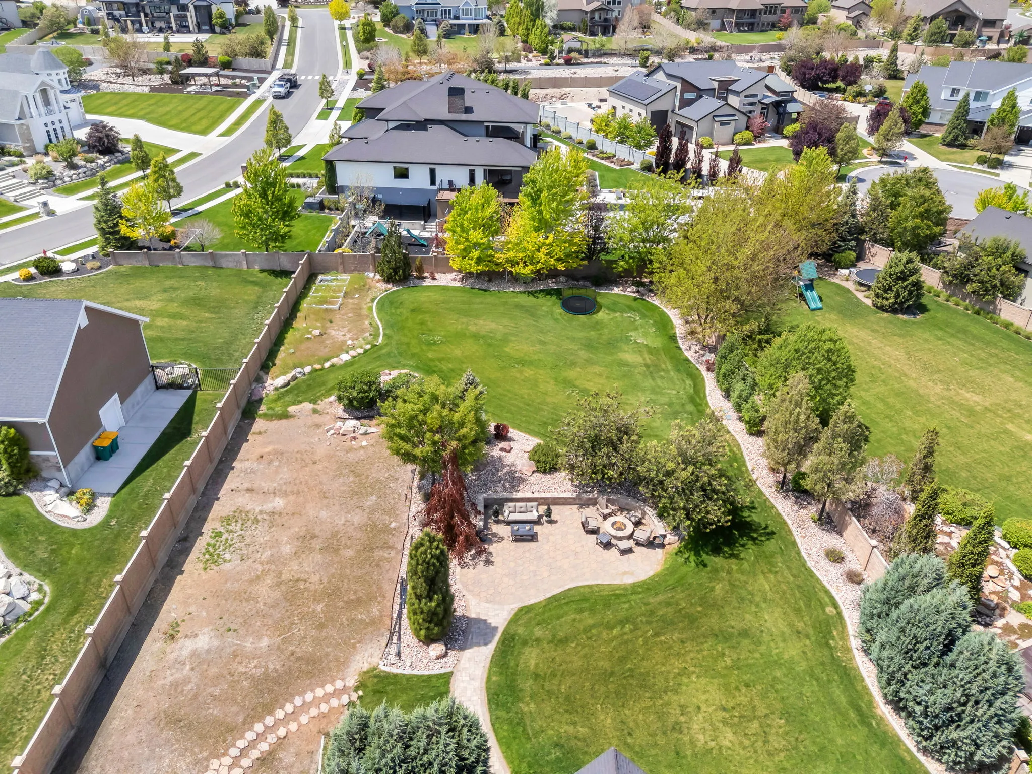 Expansive backyard featuring a stone patio with a built-in fire pit, multiple mature trees, green lawn areas, and a defined landscaping border