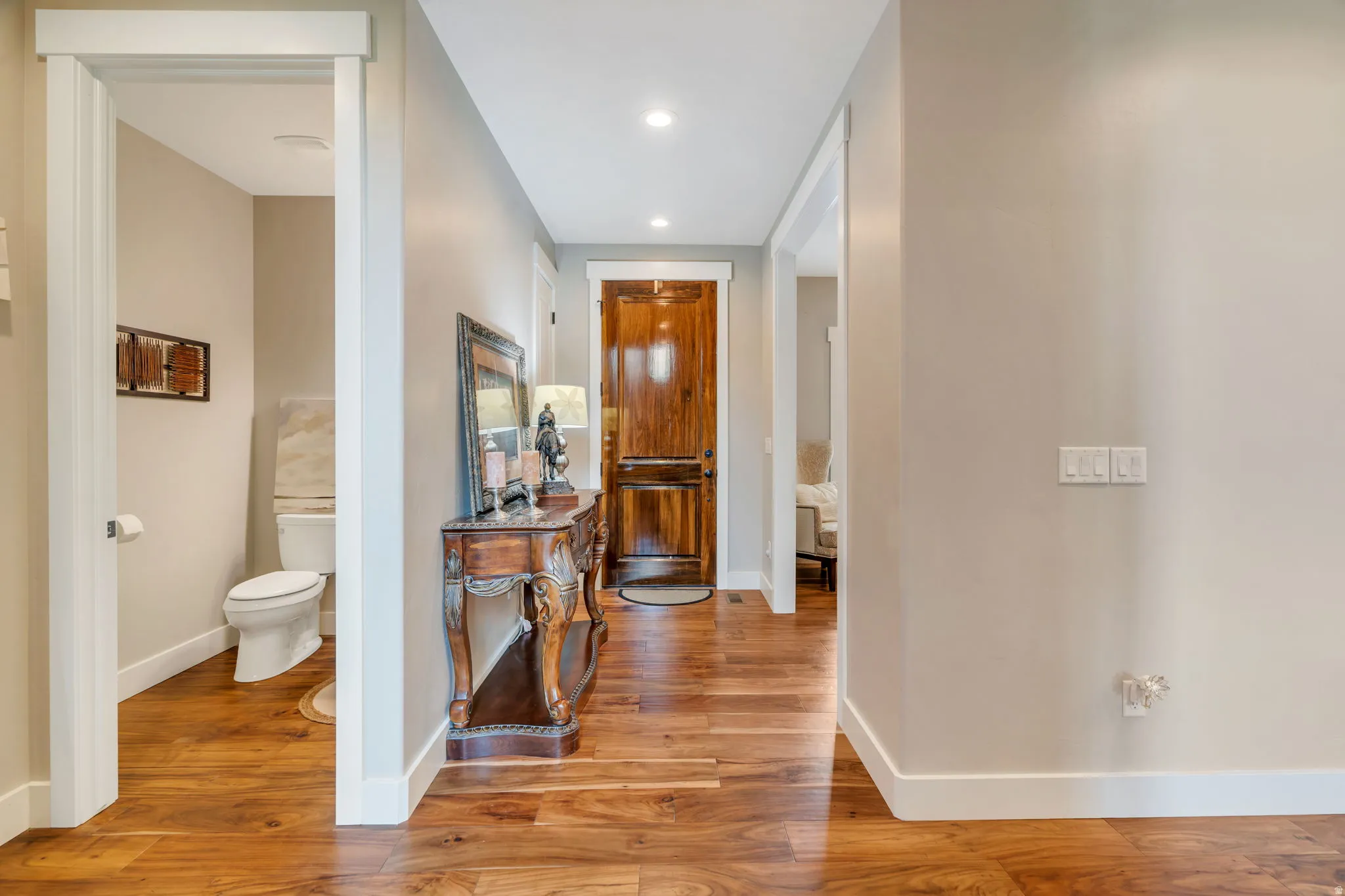 Rich wood-finish flooring extends through this interior hallway, featuring a solid wood door, recessed lighting, and a neutral wall palette