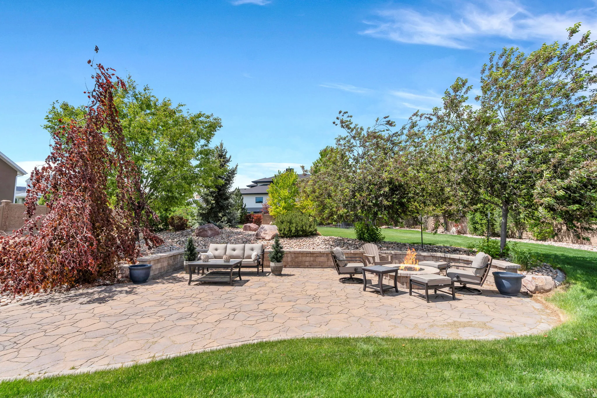 Expansive outdoor patio featuring flagstone-style pavers, a built-in fire pit, and a tiered landscaped garden with mature trees